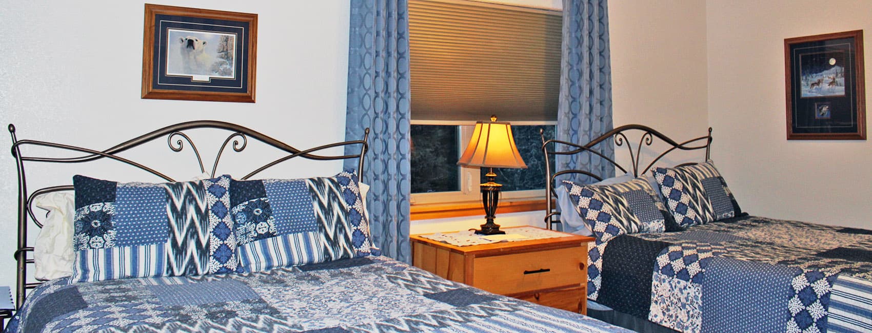 A cozy bedroom featuring two beds with blue patchwork quilts, a bedside table with a lamp, and framed artwork on the walls.