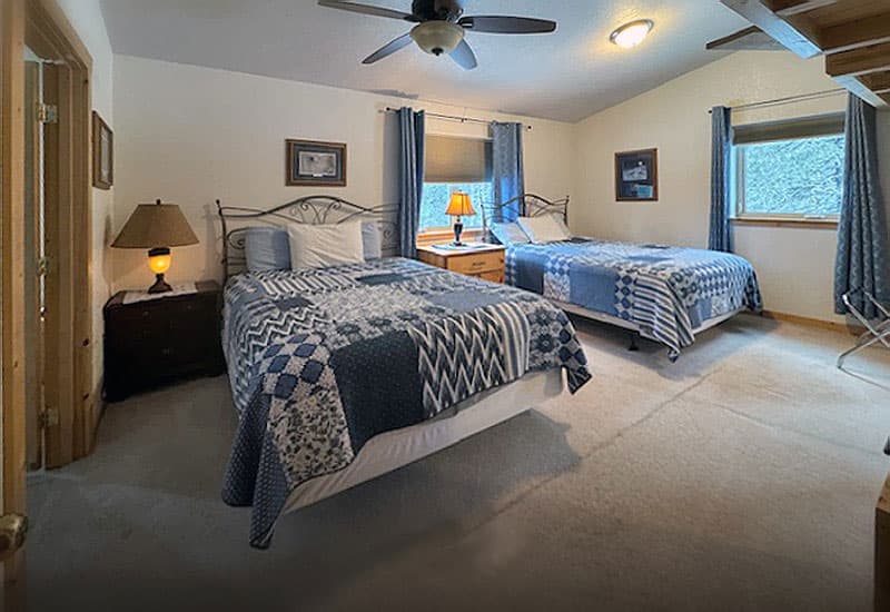 A cozy bedroom featuring two beds with patterned blankets, a lamp, and a window with blue curtains.