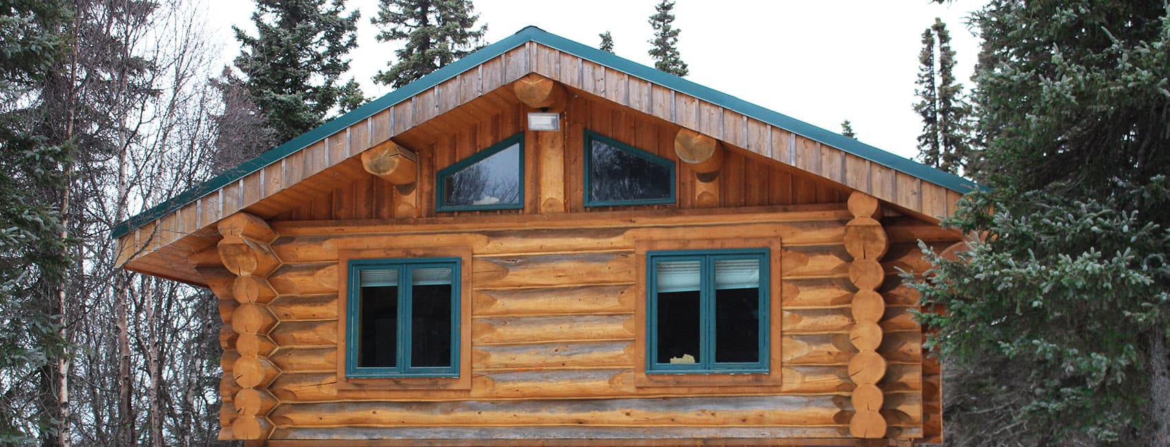 A wooden log cabin with a sloped roof and green windows, surrounded by trees.